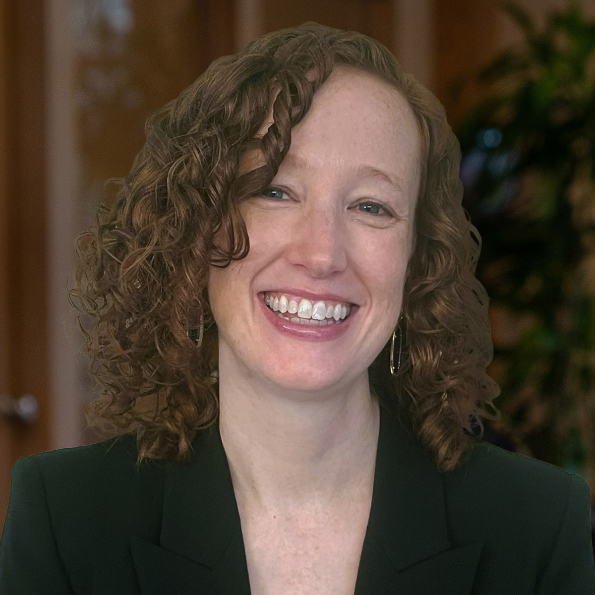 Up close headshot of a smiling woman with curly, brunette hair dressed in a blazer in front of a blurred background