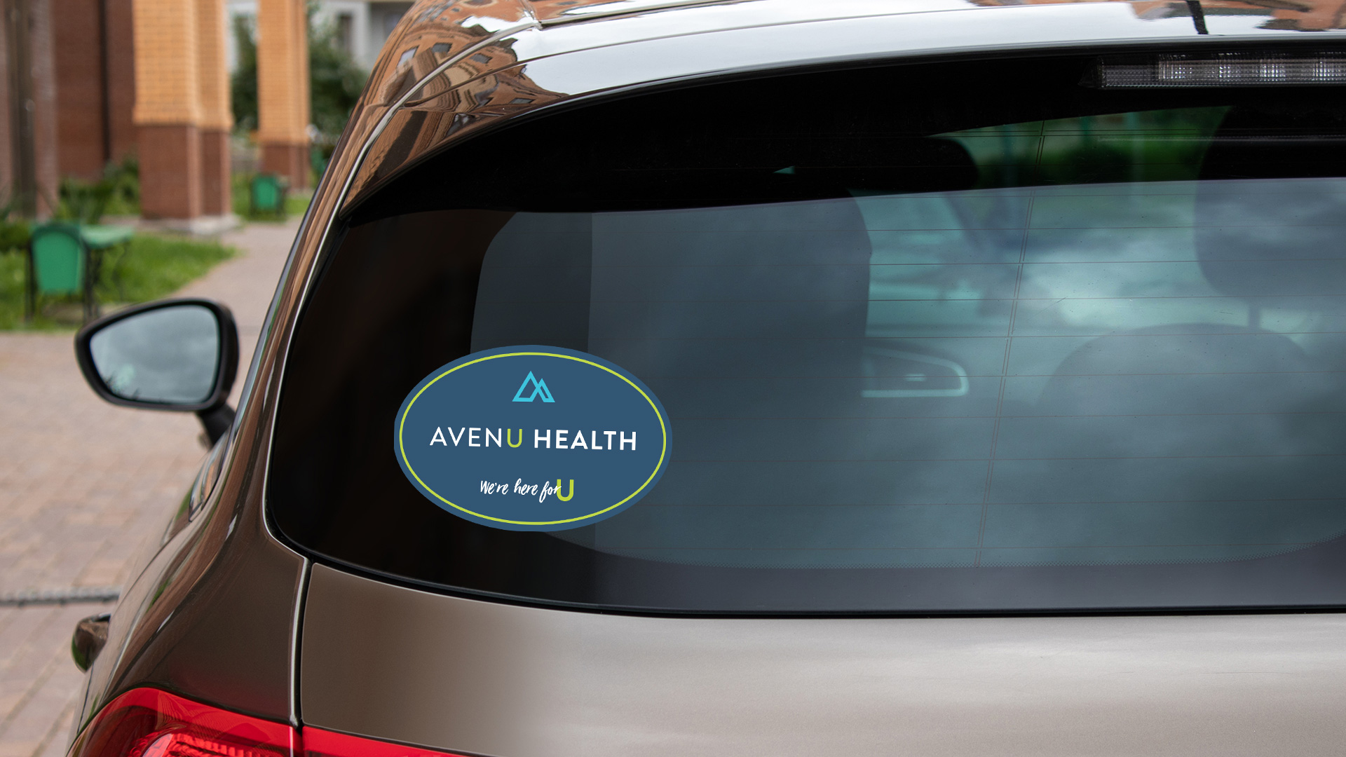 Avenu-Bumper-Sticker Navy blue oval bumper sticker on the back of a car window that reads "Avenu Health: We're here for U"