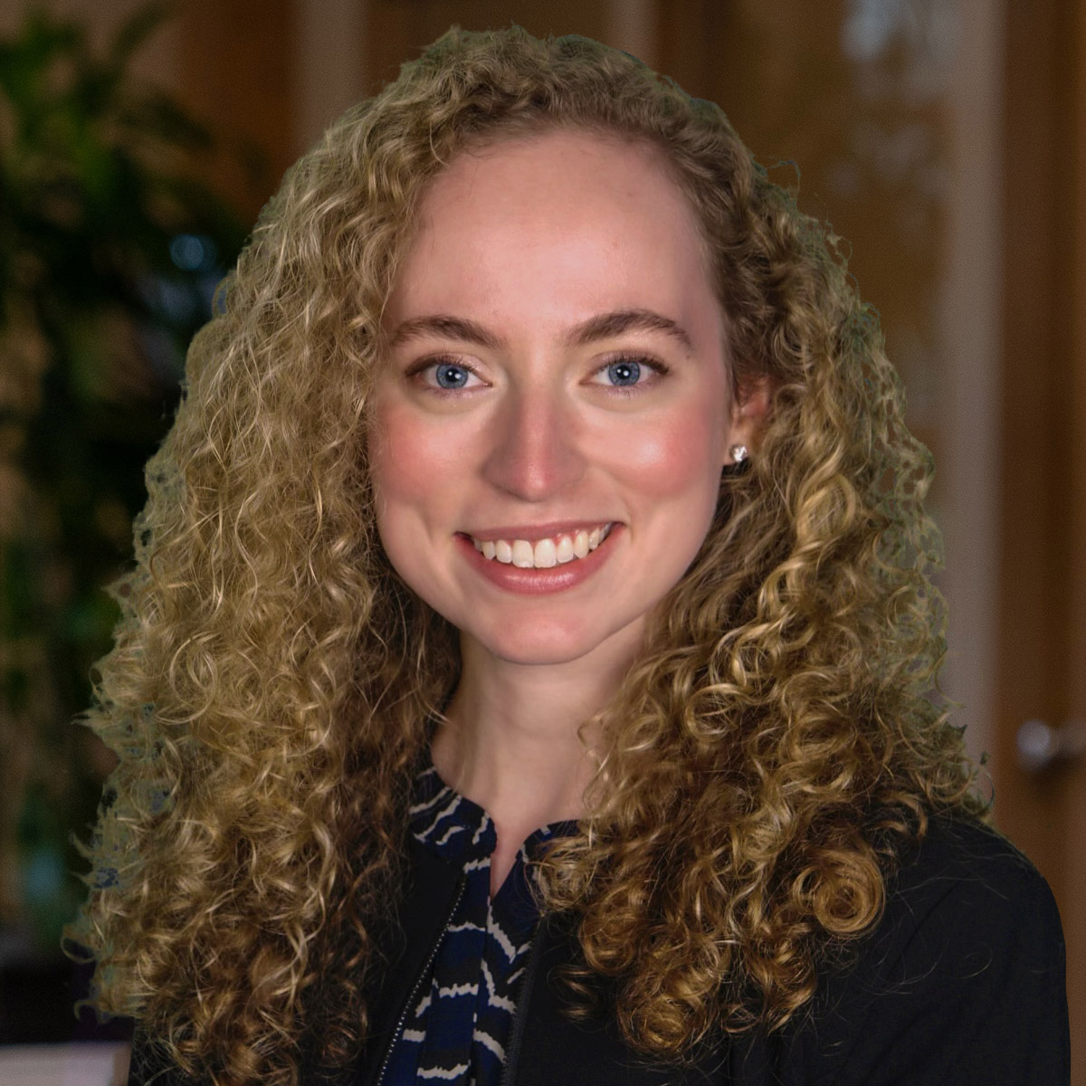 Headshot of a smiling young woman with blonde, curly hair dressed in corporate attire in front of a blurred background