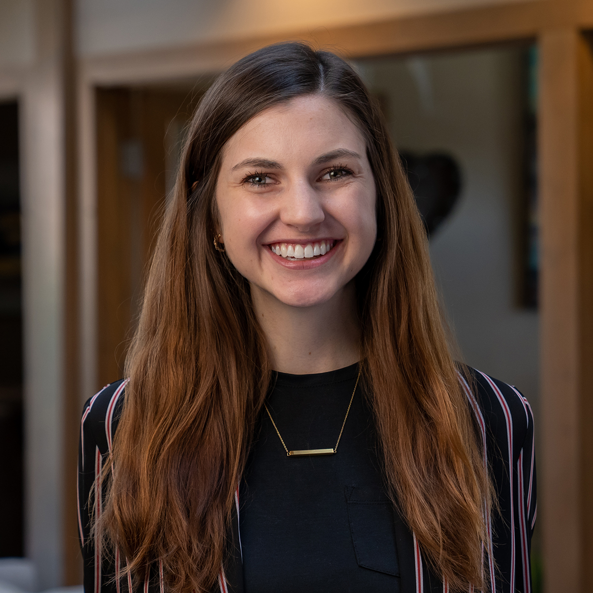 Headshot of a smiling young woman with long, brunette hair dressed in corporate attire in front of a blurred background