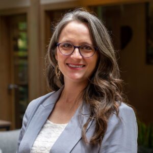 Headshot of a smiling woman with glasses and curly, brunette hair dressed in a blazer in front of a blurred background